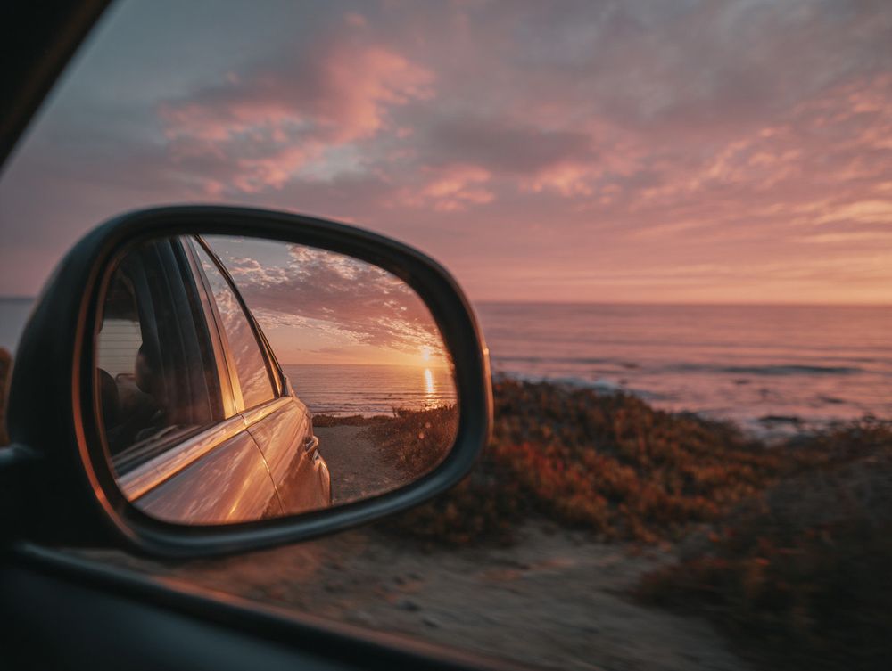Cinematic Canon EOS shot through a car window of a pastel pink sunset over a calm coastal landscape with distant sea horizon, warm golden light on clouds, realistic reflections on glass, subtle interior shadows, soft-focus background --ar 4:3 (Prompt credit: Alfonso Rosenberg / @blackowl777). (Prompt credit: Alfonso Rosenberg / @blackowl777).