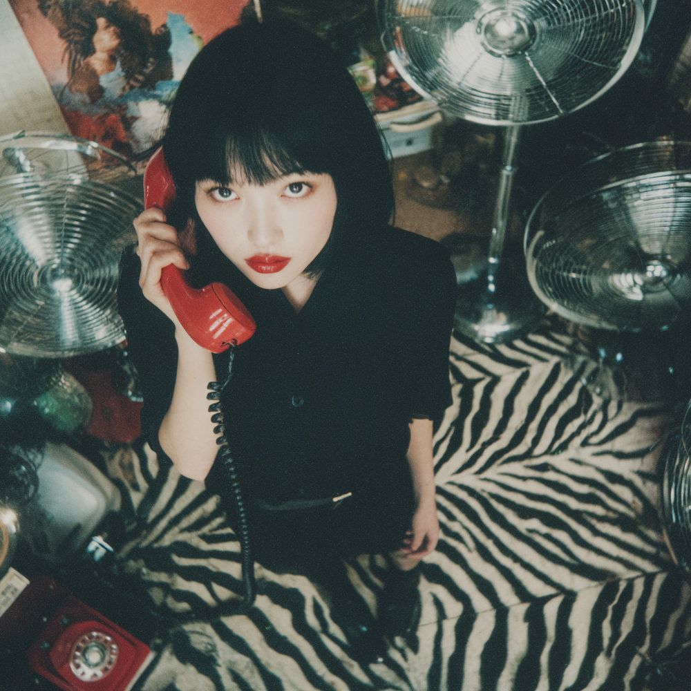 Overhead Shot This extremely grainy photo was taken with a Lomo LC-A using ISO 100 film. It features an overall bluish color cast, realistic depth of field, beautiful light and shadow, and a zebra print floor. On the right side of the frame stands a stunningly beautiful Japanese film actress wearing a black dress shirt and black jeans. Her beautiful black bob hair flows in the wind and falls around her face. She has pale skin, large, beautiful eyes, bright red lips, a sharp jawline, and a small face. She holds a red landline phone in her right hand and the receiver in her left. She tilts her head and winks at the camera. Numerous antique electric fans are installed around her. (Prompt credit: 栗橋勇 / @Dd41Giant).