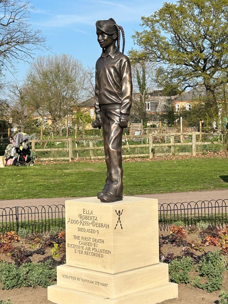 A bronze life size statue of a 9 year old girl in a park on a plinth saying Ella Roberts Adoo Kissi Debrah, died aged 9, the 1st death caused by excessive air pollution ever recorded