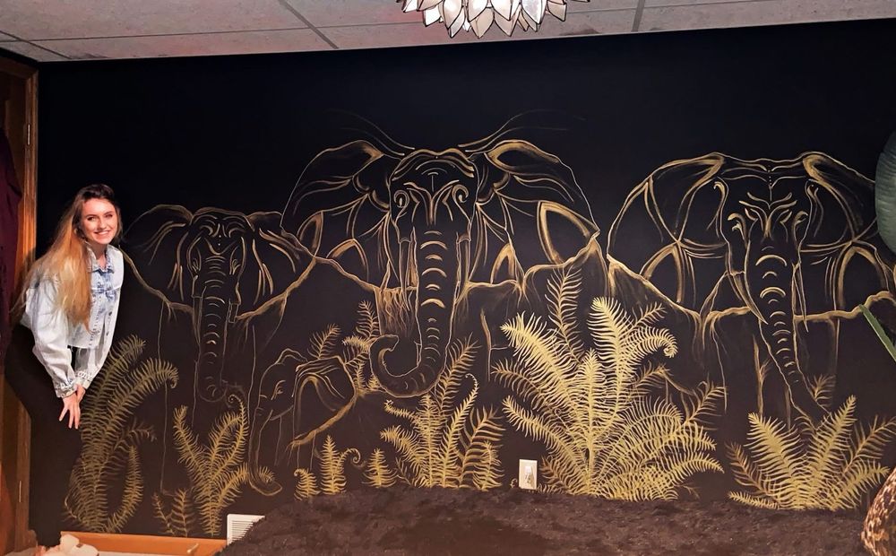 Me, posing to the left of a large scale golden elephant mural against a black painted wall in a bedroom. Three elephants march towards you, with a tiny baby elephant slightly concealed in the tall fern fronds that line the base. 