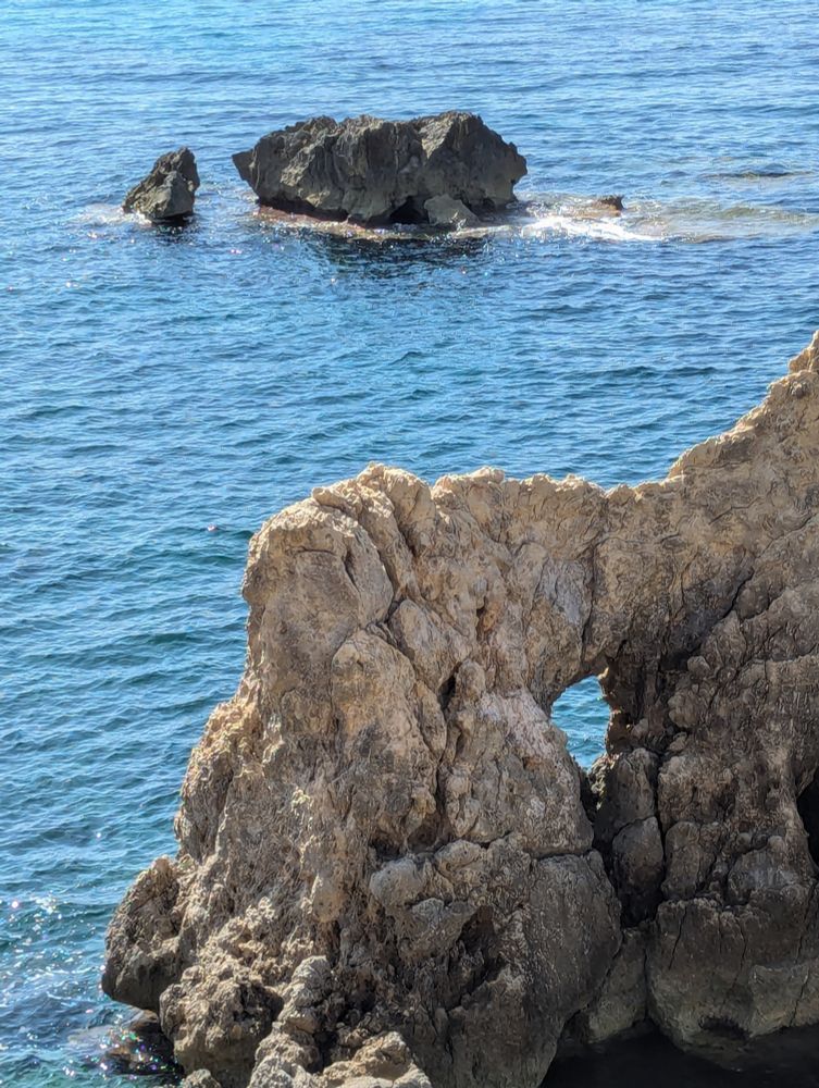 Turquoise blue sea and a cliff at the front with a whole through which the bright sea is visible. Two dark, angular grey rocks in the distance, one large, the other much smaller, and a tiny one to the right of them.
