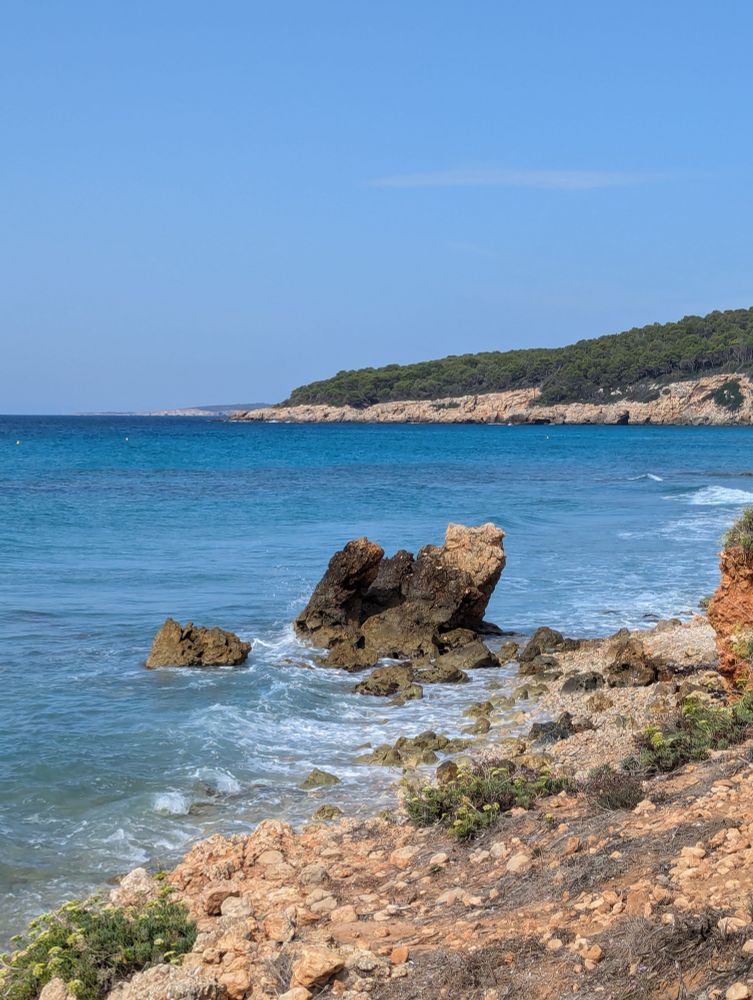 Blue bay with rock immersed in water, and rocky shore at the front. Pine trees in the distance at the opposite shoreline.