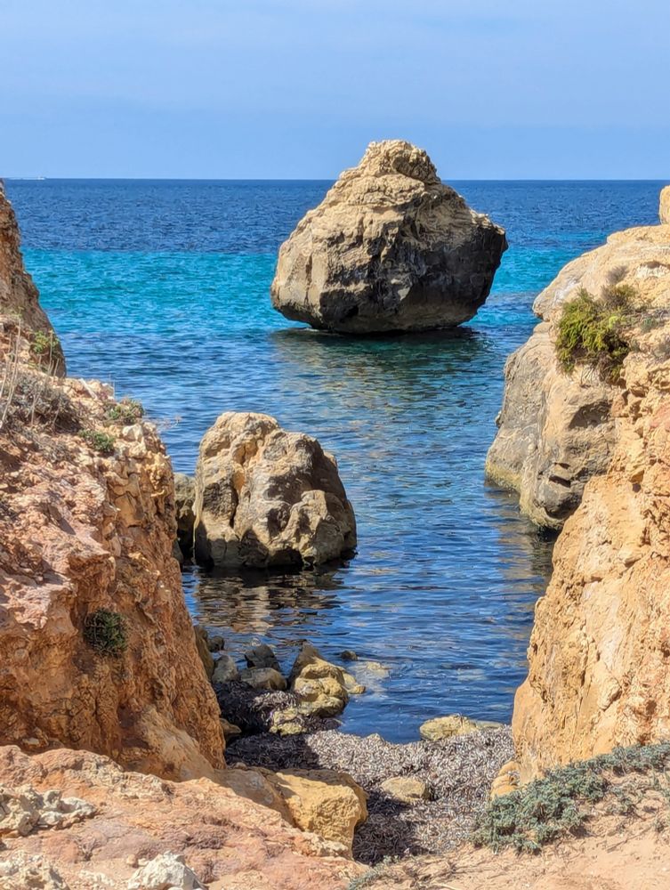 Blue turquoise-blue sea with a large triangular, rounded rock in the sea in the upper middle area. A smaller rock at the lower left side.  Sand-coloured rocks rise to both sides in warm sunshine.