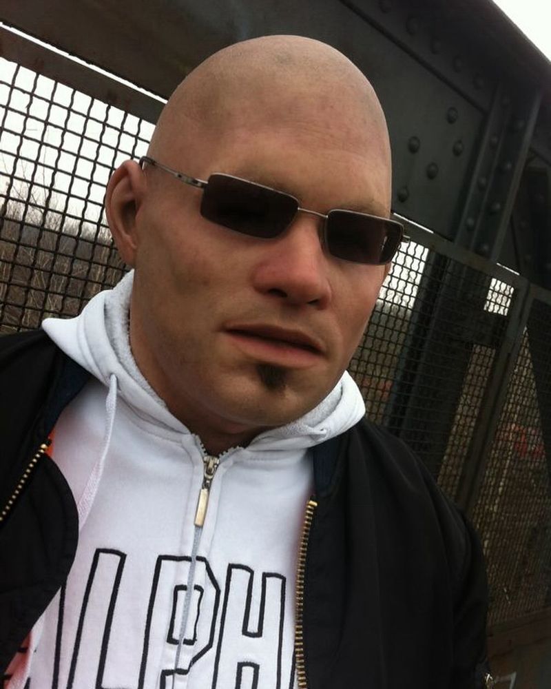 Bald man with sunglasses and a goatee wearing a white hoodie with the word "ALPHA" and a black jacket, standing in front of a metal fence.