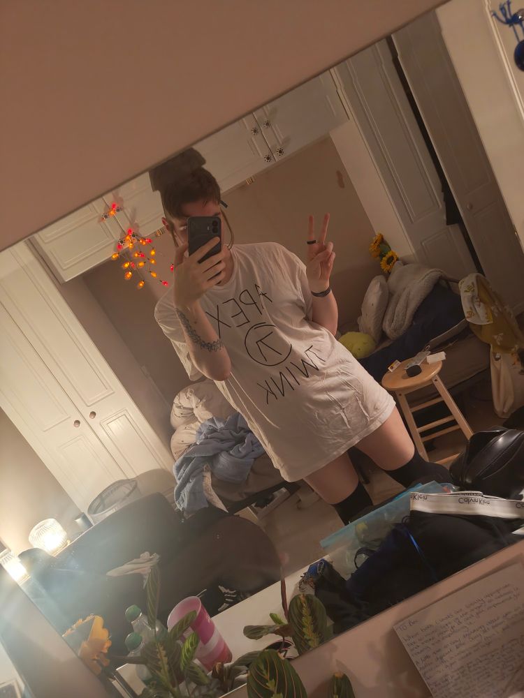 A bedroom mirror selfie of a transfem person making a peace sign gesture. They are wearing a baggy white XXL T-Shirt which reads "APEX TWINK" and black thigh-high socks.