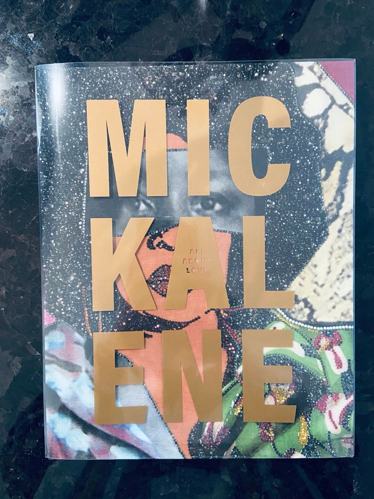 A photograph of a large book sitting on a black stone countertop. The book has a clear acetate dust jacket on which the name “Mickalene” is printed in large sans serif bronze-gold letters. The name is broken into three parts of three letters and spread over three lines. The words “ALL ABOUT LOVE” are cut out of the letter A. Behind the cover, an image of a collage of a beautiful, luminescent Black woman’s face covers the book. Her eyes are taken from and black and white photograph. Her hair, jacket, nose and lips sparkle with sequins.