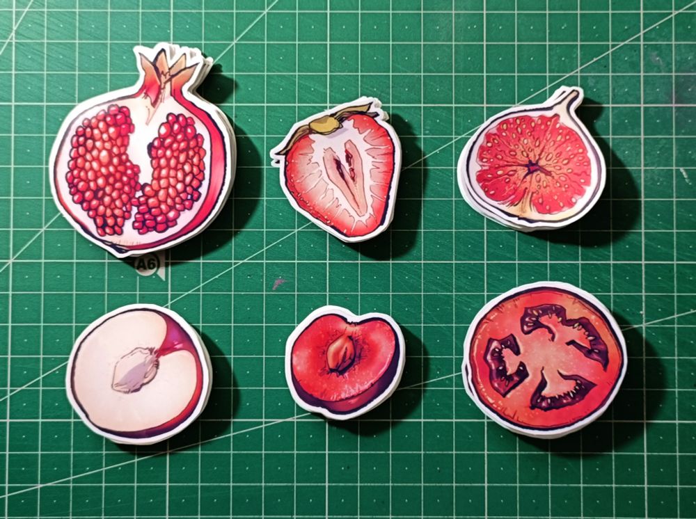 Photo of a green cutting mat with small stacks of stickers on top of it. There are 6 different sticker designs, all of them of a different red fruit cut in half. From top left to bottom right: a pomegranate, a strawberry, a fig, a plum, a cherry and a tomato.