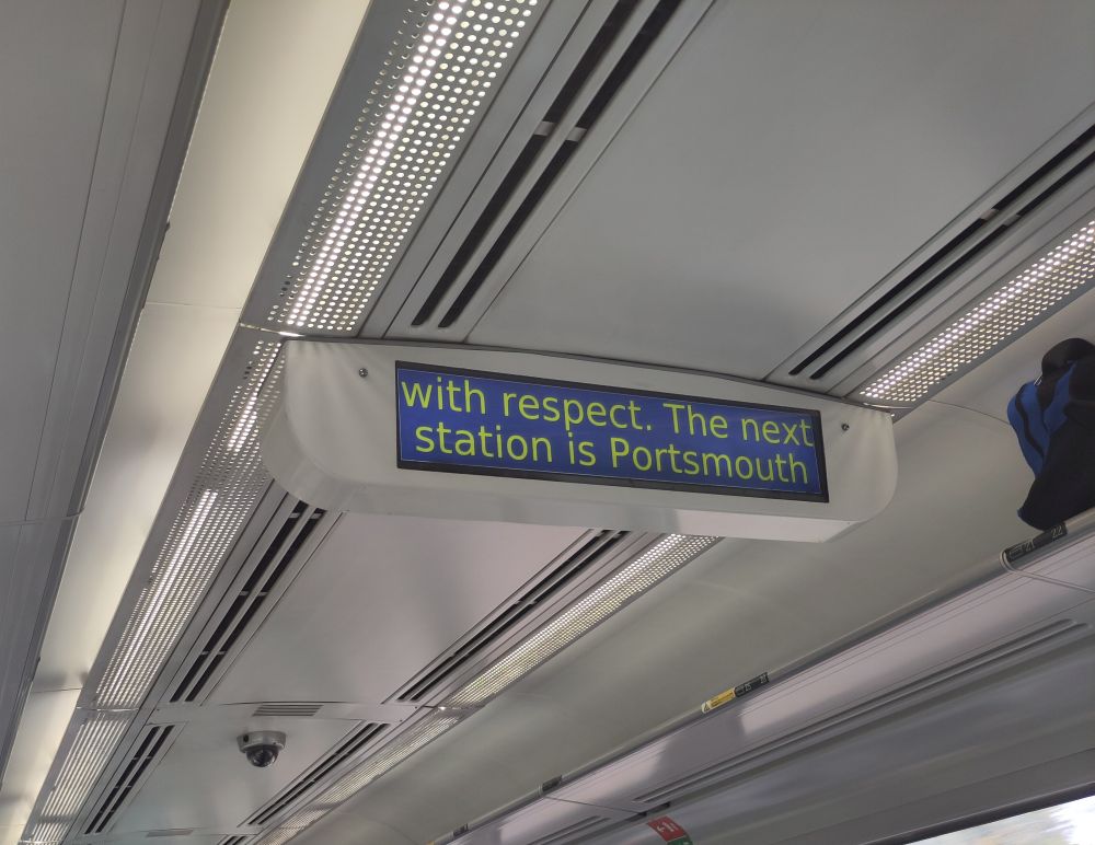 A digital display screen inside a train shows the message: "with respect. The next station is Portsmouth" in yellow and green text on a blue background. The display is mounted on the ceiling of the train carriage, surrounded by metal panels and overhead lights.

