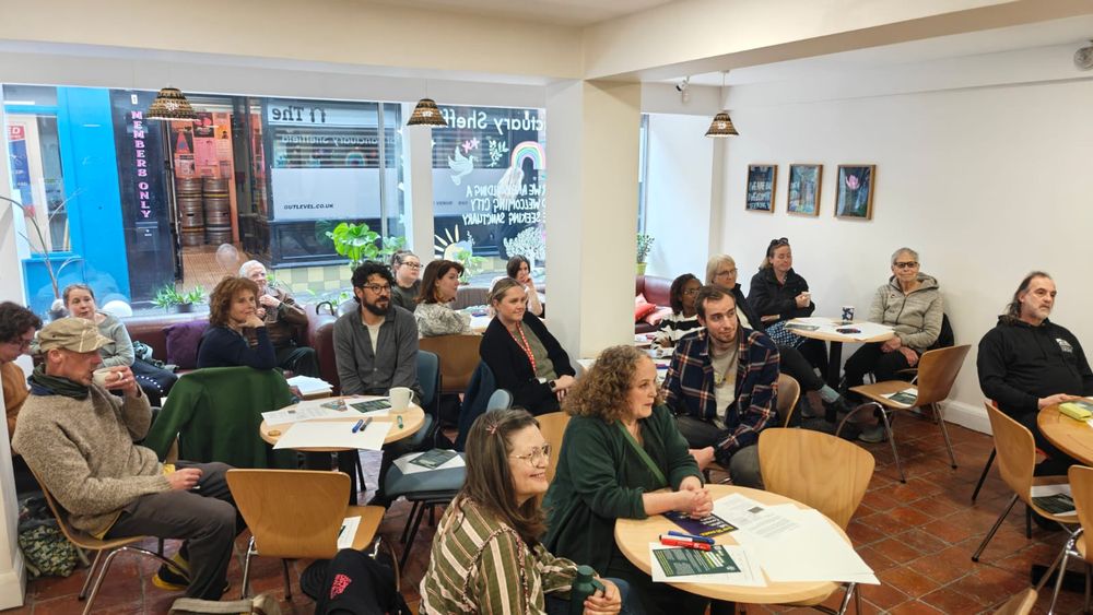 A photo of a small room full of people, sat 'cabaret style' and listening to a speaker out of shot