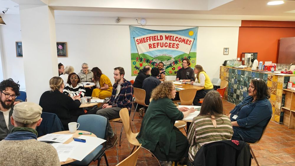 A photo of a small room full of people, sat 'cabaret style' and speaking with each other in front of a banner on the wall with the words "SHEFFIELD WELCOMES REFUGEES"