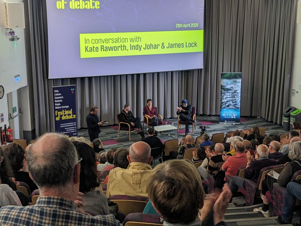 A photo of three people sitting on a stage in conversation with each other, in front of a screen with the words "In conversation with Kate Raworth, Indy Johar & James Lock"