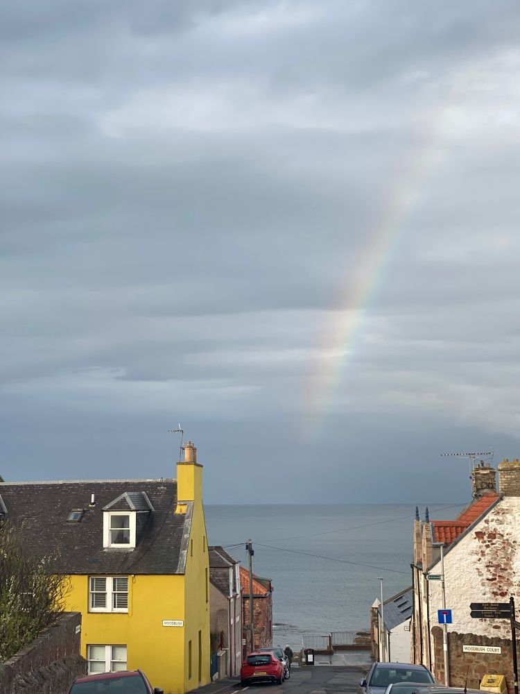 A street leading to the sea, with a yellow house on one side, a white one on the other, and beyond them, in the ocean and overhead, a rainbow. 