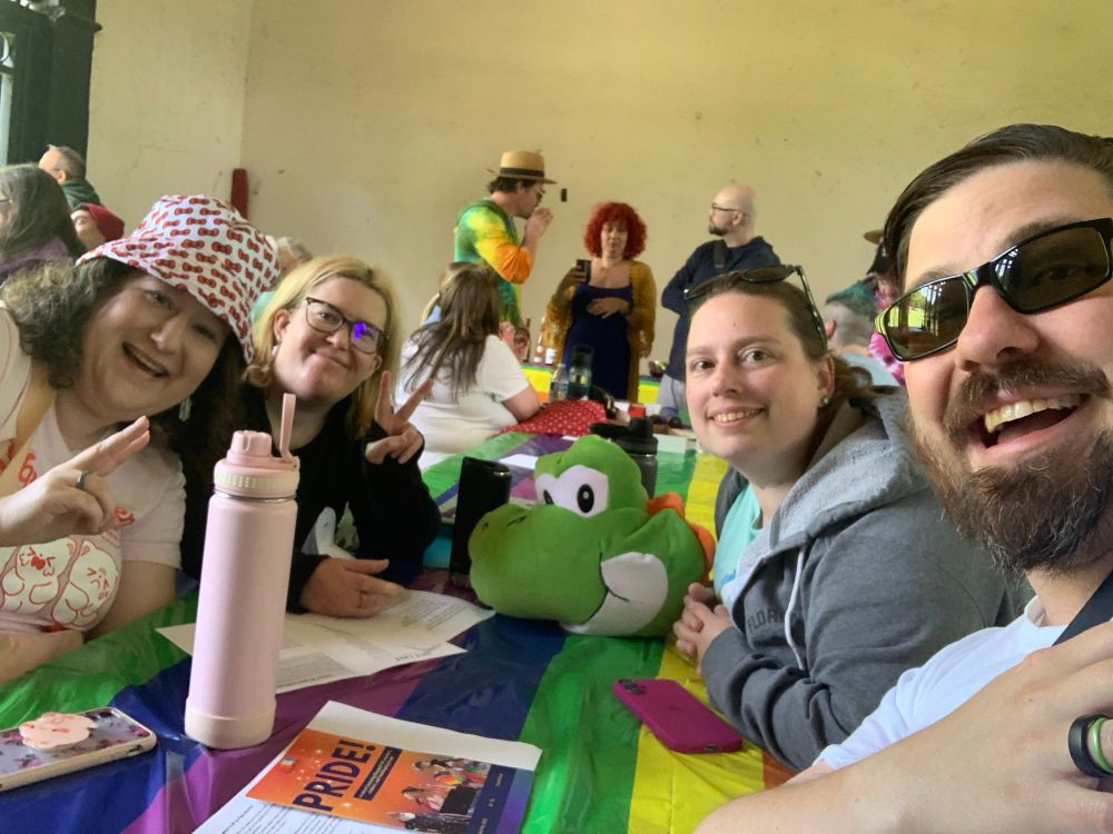 A photo of me and three of my friends leaning in and smiling at a picnic table covered in a rainbow-striped plastic cover.  The friend next to me and I are making peace signs with our fingers. 

There are programs and lyric sheets on the table, as well as a small pillow shaped like Yoshi’s head, a couple of thermoses, and a couple of phones.