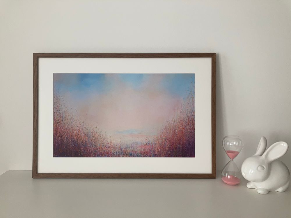 A framed print standing in a white surface. Next to it is an hourglass with baby pink sand, and a white soft ceramic bunny.
The print is of a painting with big soft pink clouds and lots and lots of thin vegetation along the bottom and prominently in both lower corners. Up close it looks wild but from a distance it’s a calm scene.
The print is framed in a dark wooden frame with a wide white passepartout.
