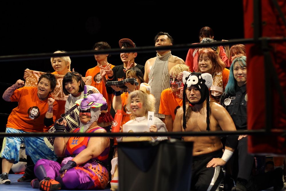 at the end of one of Kaori Yoneyama's YMZ wrestling events, the wrestlers pose together for photos. in the front row is iconic pro wrestler Aja Kong, dressed partially as Evangelion Unit-01. next to her is famously un-retired pro wrestler Kaori Yoneyama, dressed as a cake to celebrate a wrestling anniversary. the dozen or so people around them are dressed as an Angel from Evangelion and the cop from Terminator 2, in addition to normal clothes. A sweater pervert and mixed martial artist, Hikaru Sato, is also present.