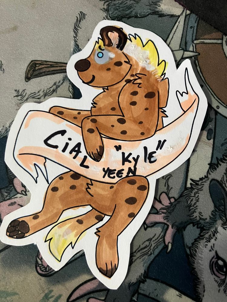 its a cute lil hyena printed and cut out on nice card stock, holding a banner. the linework is maul's. i colored it in kinda hastily to be a spotted hyena w blonde hair and a silver spot on his eye. the banner text awkwardly reads "Cial (to be pronounced "kyle") yeen"