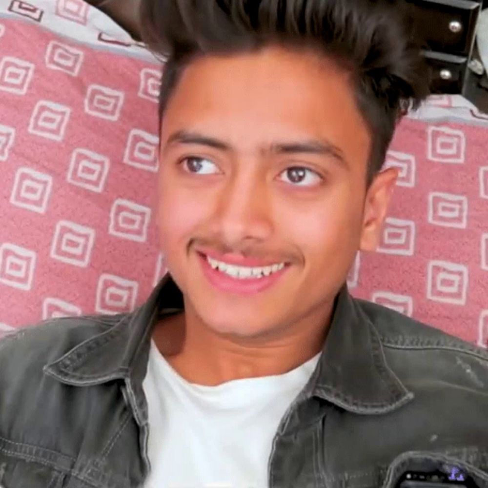 a young man with a mustache is smiling while sitting on a bed with a pink blanket .