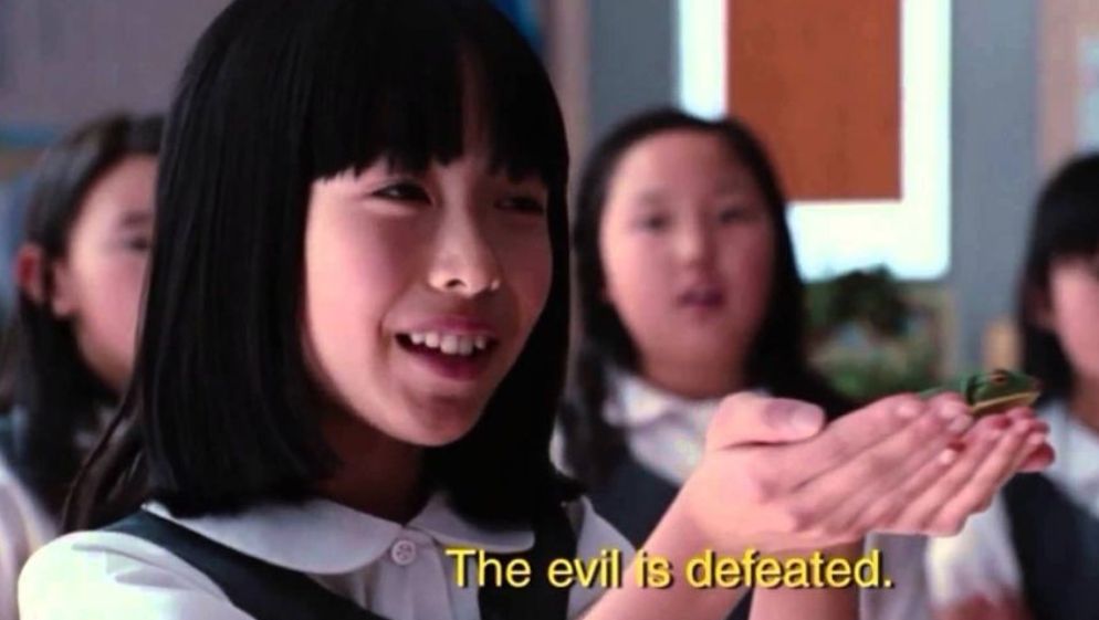 A screenshot from the horror comedy movie The Cabin in the Woods, where a young Japanese schoolgirl is happily holding a frog in her hands while saying "The evil is defeated."