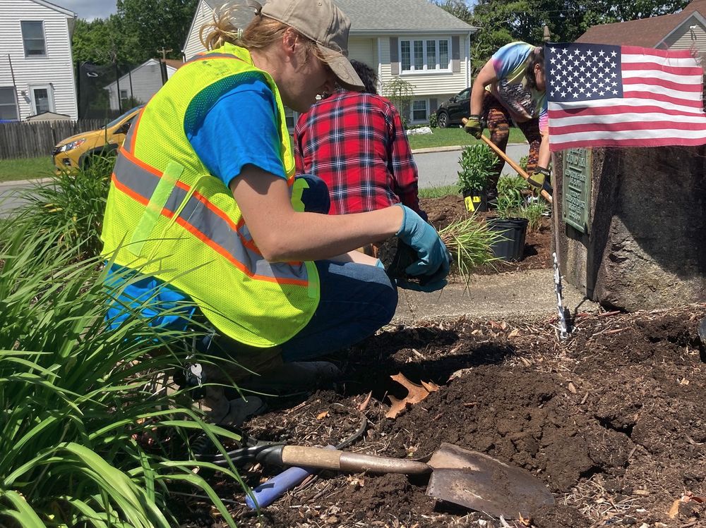 Three people are gardening near a large stone with a plaque in a residential area. One person in a high-visibility vest and blue gloves is planting grass, another in a plaid shirt is kneeling nearby, and a third in a blue shirt with a shovel is digging. Houses and a parked car are visible in the background.