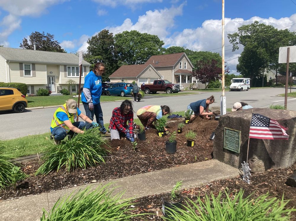 A group of people is gardening around a large stone with a plaque in a residential neighborhood. Some wear blue "EPIC" shirts, while others are in casual clothing, planting small plants from pots and digging in the soil. Parked cars, including a yellow car and a red pickup truck, line the street, with houses and a mail truck in the background under a partly cloudy sky.