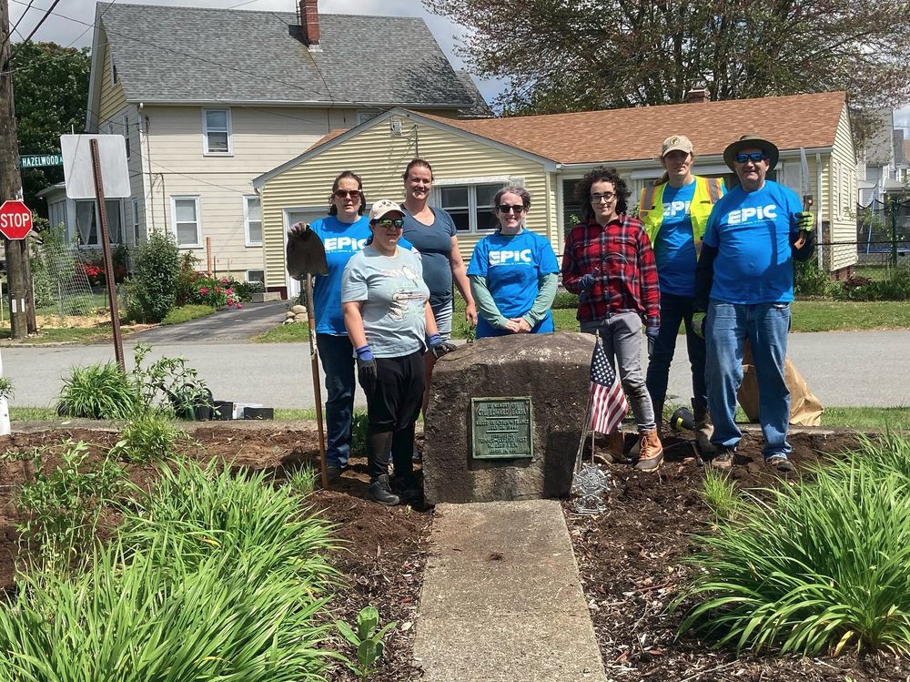 A group of seven people stands around a large stone with a plaque in a garden area near a residential street. The plaque reads "In Memory of Cpl. Edward Farina, United States Army" A small American flag and a decorative pinwheel are placed beside the stone. The group is dressed in casual clothing, with some wearing blue "EPIC" T-shirts and others in work gear, holding gardening tools like shovels. They appear to be participating in a community gardening or memorial project. In the background, there are houses, a stop sign, and a street sign for Hazelwood Ave.