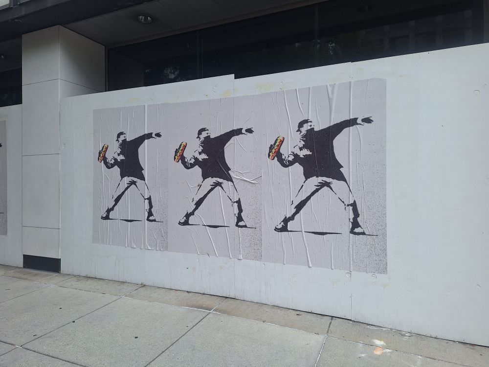 Photo of wheat pasted street art posters on boarded up vacant office building facade. Image on posters is Banksy's flower throwing stencil holding a long sub sandwich in honor of the city's U street mascot. 