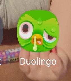 Screen grab of the Duolingo app icon where the green owl's face is contorted, one eye sagging, sweating like a pig, and a giant booger trailing down from its beak/nose. Background image is of an upside down toddler arm holding a toothbrush.