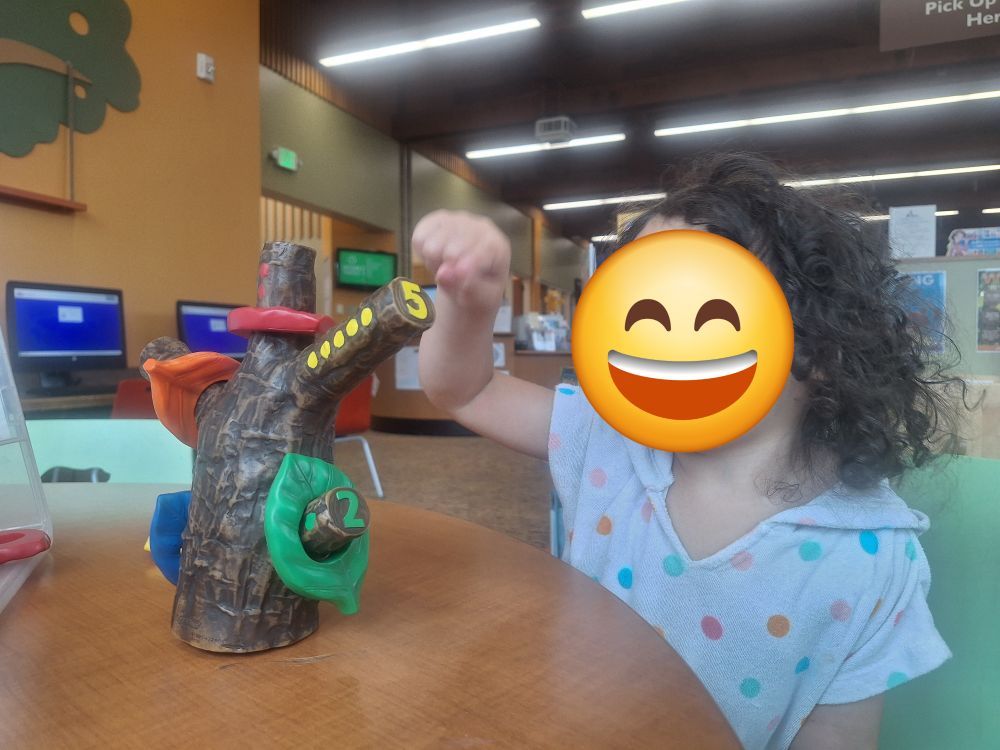 Photo of a young toddler girl with unruly curls and a white hoodie shirt with colorful polka dots plays at a children's table in a public library. She is pointing to the counting tree toy, specifically the yellow 5 branch that is just waiting for her to count out the yellow leaves. There is a joyful smile emoji 😄 over her face for privacy.