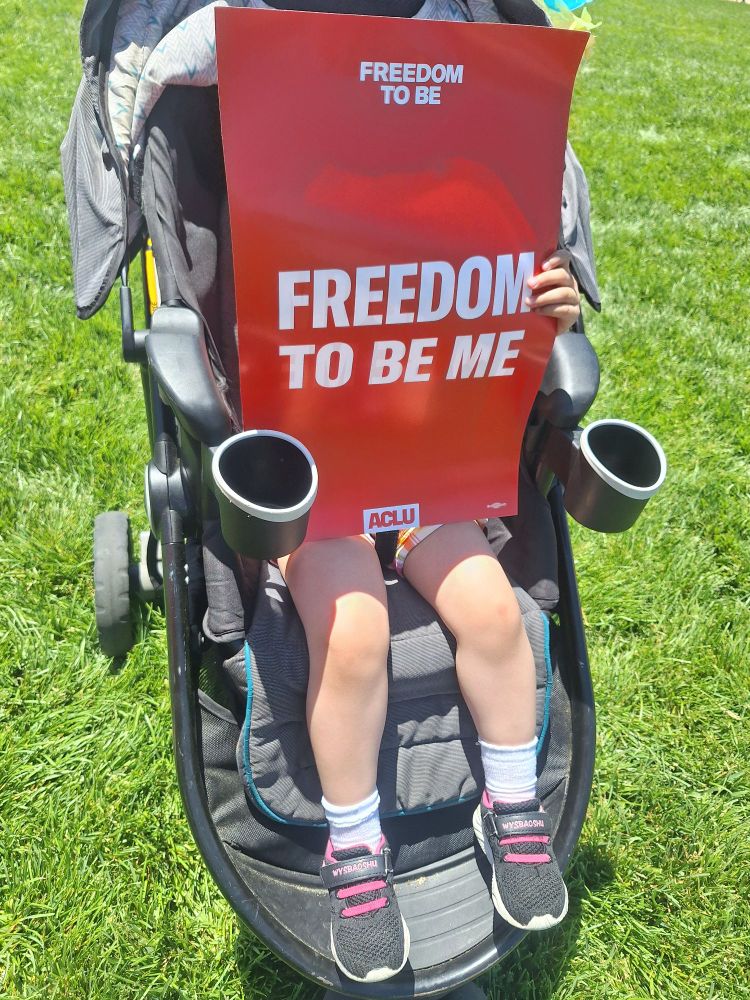 Stroller in a lush grass lawn and with two toddler legs with a bit of plaid shorts showing and wearing white socks and black and pink sneakers, toddler is holding a red sign in front of them that says FREEDOM TO BE and FREEDOM TO BE ME in white. At bottom of the sign is a whit box with ACLU in red.