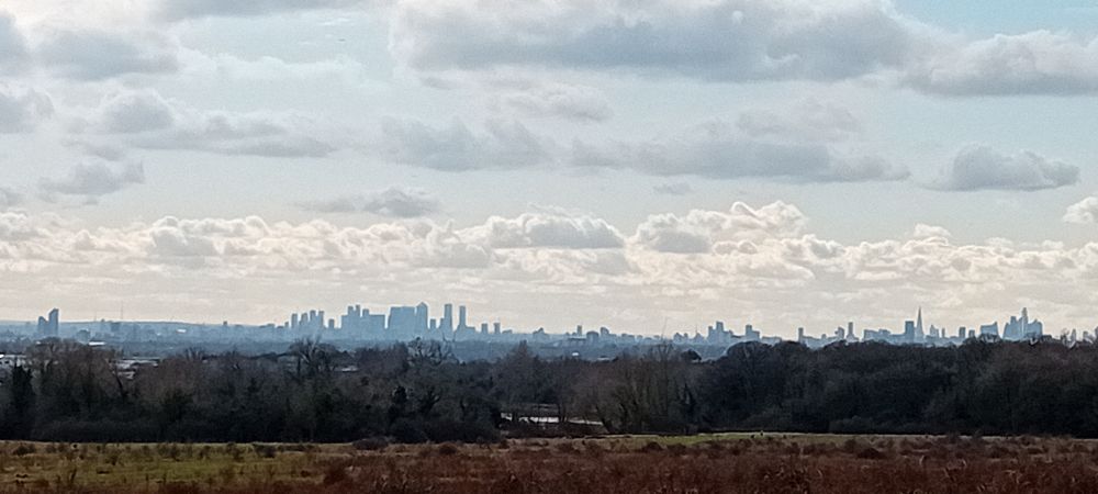 Wide-angle panoramic shot.
Mostly sky, high clouds, backlit by a low sun. In the foreground, scrubby heath, then trees, then on a distant, hazy blue horizon the silhouettes of Canary Wharf and the City of London.
In the expanse of sky, a speck of a skylark…you had to be there, but, oh, the song!
From Beacon Hill, Hainault Forest Country Park, 3 March 2024