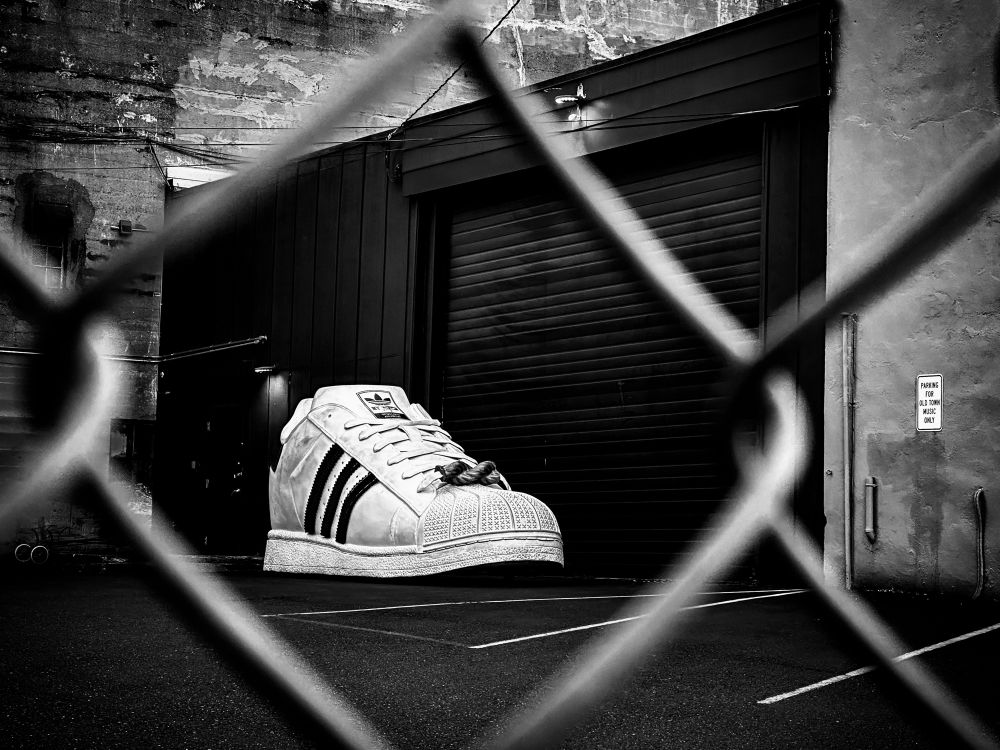A black and white photo of a giant sized adidas shoe sitting in a parking lot behind a building. The photo was taken through a chain link fence and framed in one of the links.