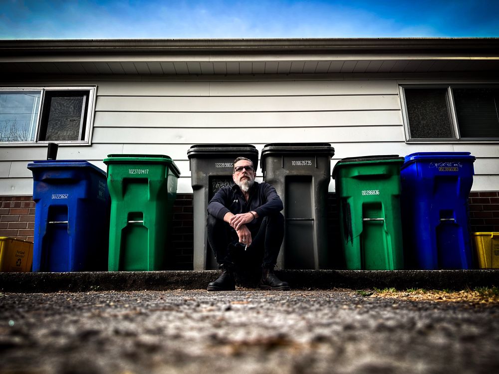 Color self portrait of me sitting on a curb in a parking lot with 8 trash bins behind me.
