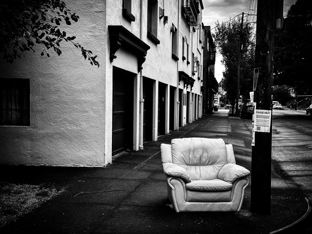 A black and white photo of a puffy white leather chair sitting on a street corner.