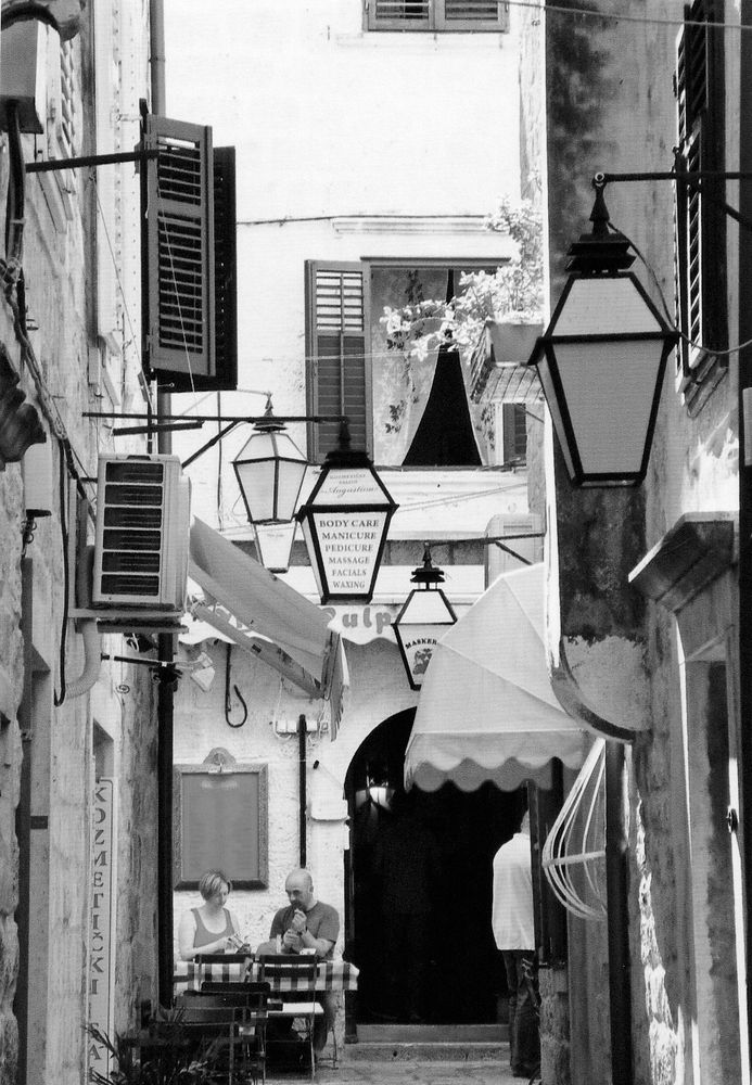 At the end of a narrow street, a couple are sitting at an outside table eating lunch. The homes and shops on the street have awnings, shutters and large outdoor lights mounted on the walls. The cafe has chequered table cloths.