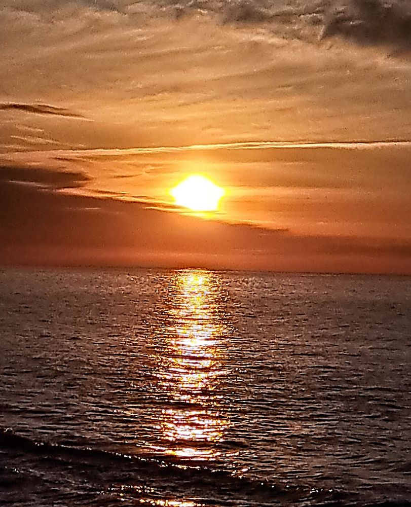 The sun rises above the clouds on the horizon. It shines right across the water, to the lake shore. The sky is shades of orange and grey.
