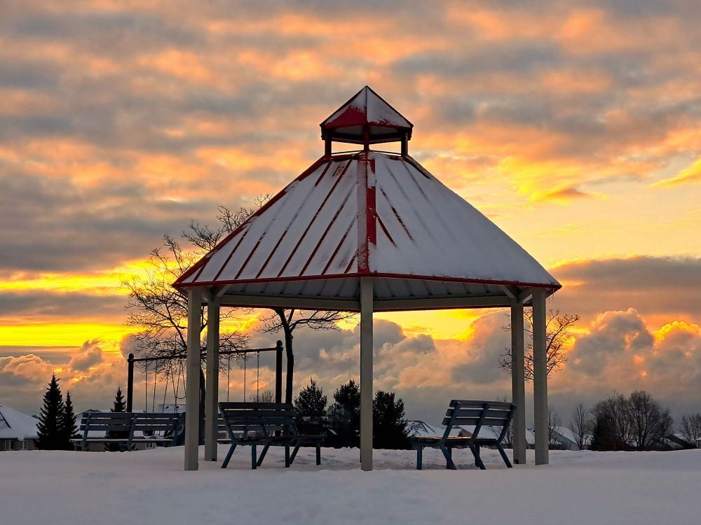 A winter scene with snow on the ground. A red gazebo has 2 benches underneath it.  The sun is just rising above the low cloud cover and the sky above is shades of gold, orange and grey.