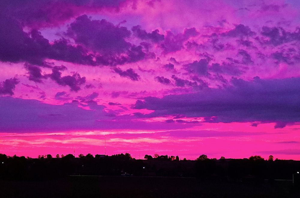 At the bottom of the photo is the dark silhouette of farmland and trees.  On the horizon, the sky at sunrise is a bright pink and above that, the sky is a mix of pink, mauve and purple.