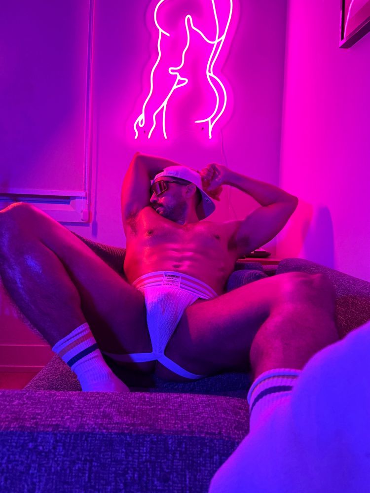 Muscle stud lies back on couch in jockstrap and crew socks, sniffing his pits, his abs illuminated by a pink neon sign of a male physique. 