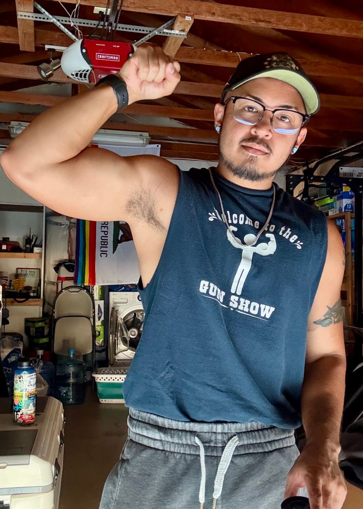Selfie in a cut off shirt that says “Welcome to the GUN SHOW” with left bicep flexed