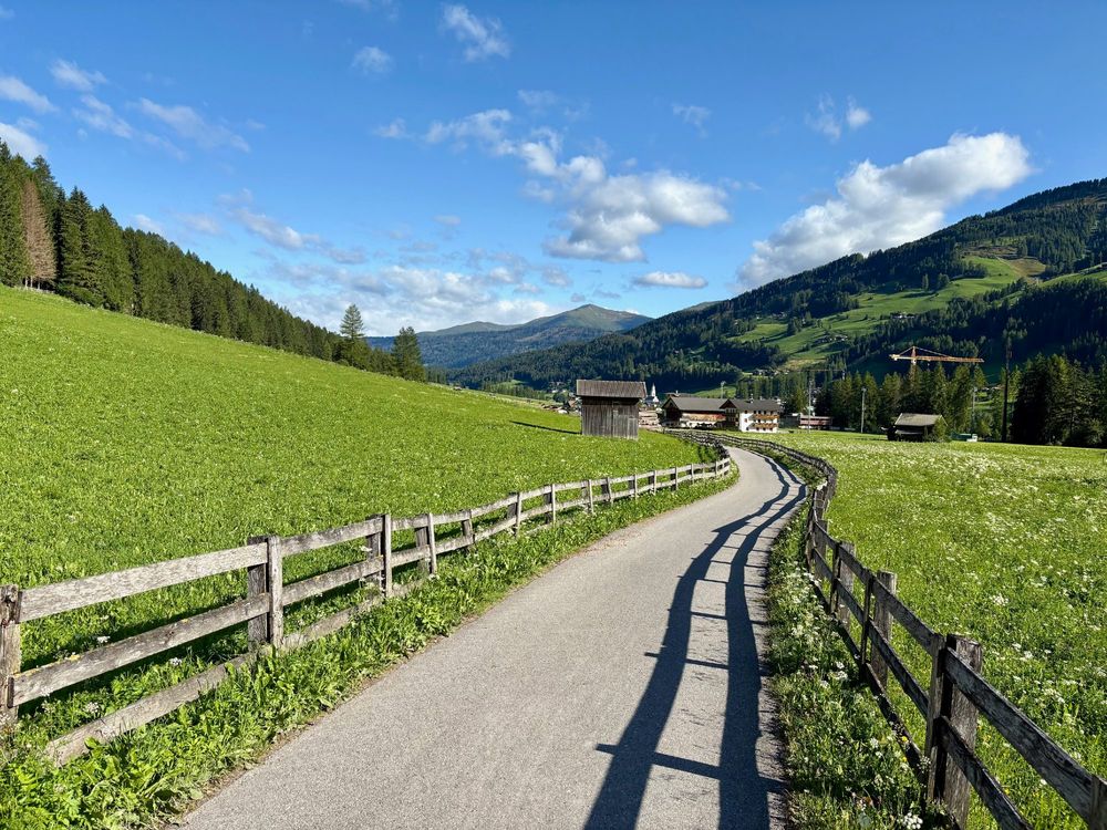 A path through a green valley surrounded by mountains. A blue sky with a few white clouds highlights the clear weather when the photo was taken.