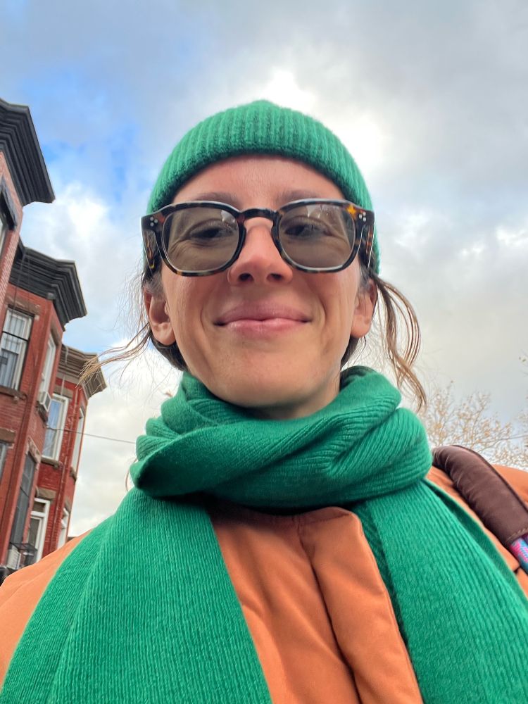 Tyler a white brunette female smiling with a beanie, scarf and sunglasses. It’s sunny and cloudy.