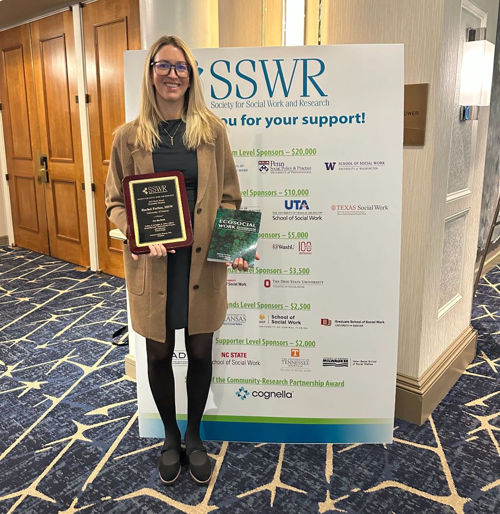 GSSW's Rachel Forbes stands in front of an "Society for Social Work and Research" sign, holding a plaque and a book in each hand.