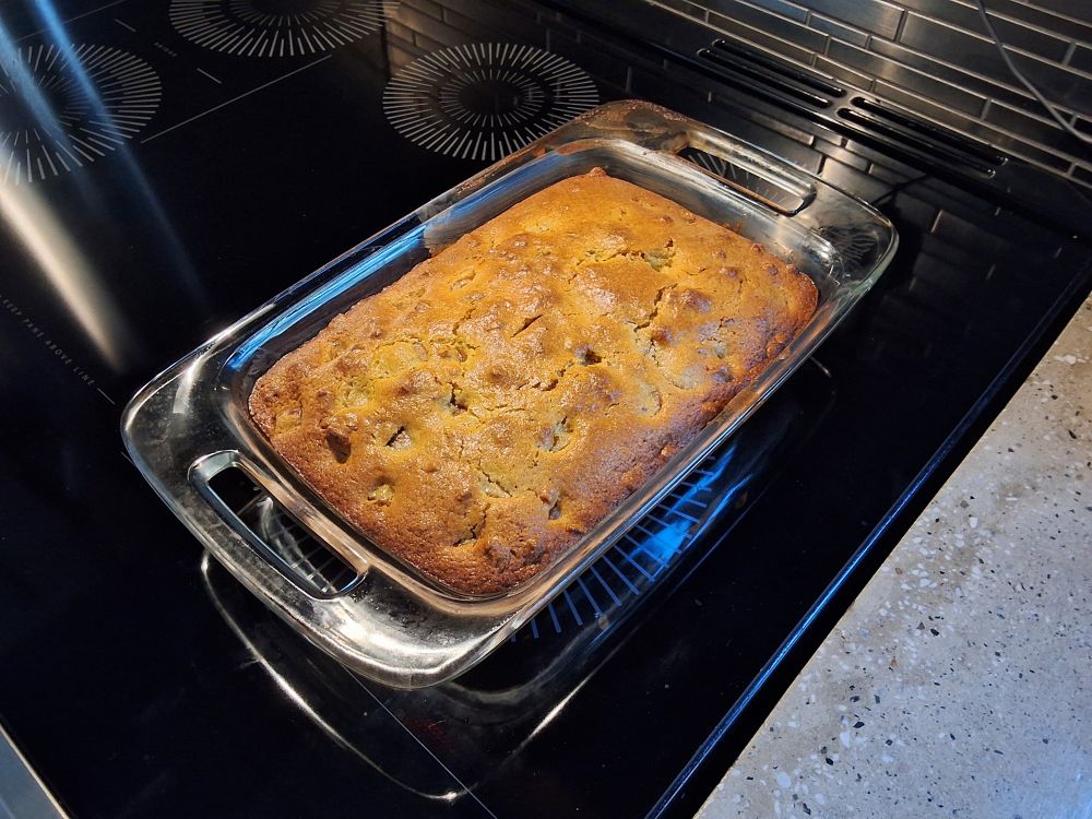 A fresh baked pan of cornbread in a clear glass baking pan sits on top of a black glass stove top. The bread is a rich looking yellow brown with variegated darker brown spots across the top and around the edges