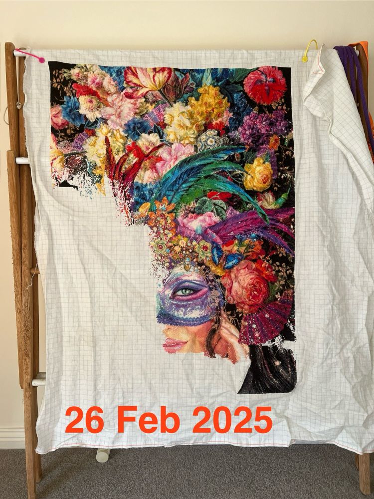 Unfinished cross stitch. Design includes half of a masked woman’s face with a head dress of flowers and feathers.