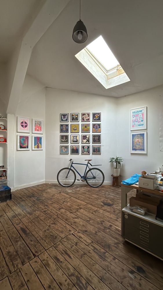 Photo of a studio room with worden floor, white walls with many framed Illustration and a ceiling window.