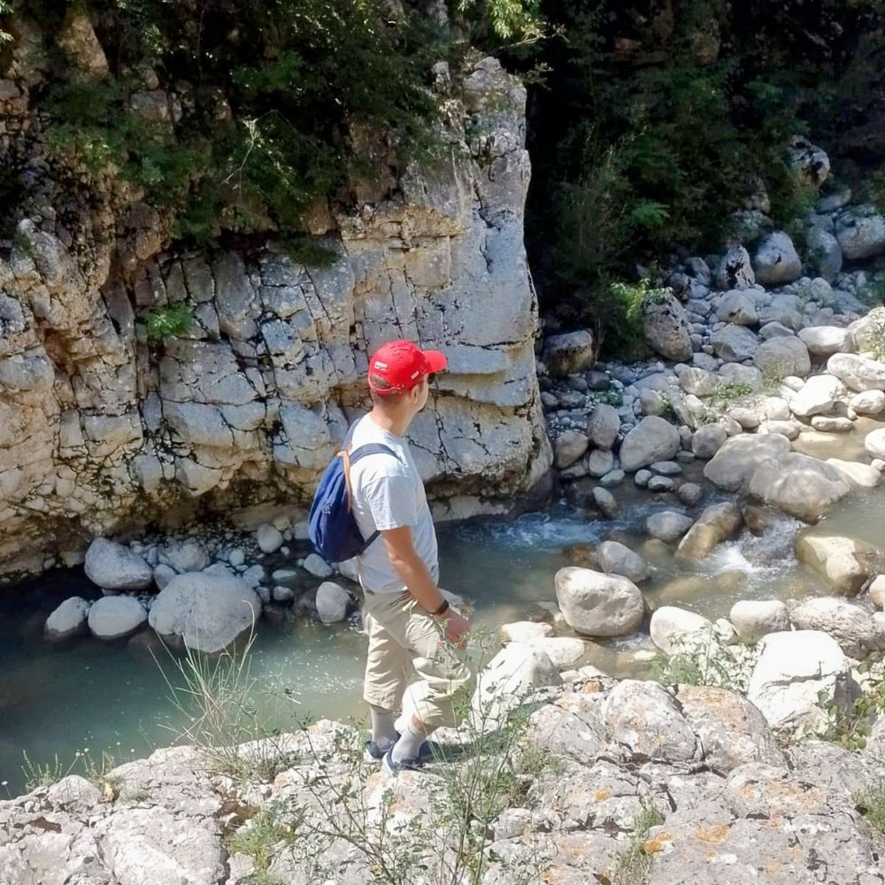 Photo of a white man with a red hat and a blue backpack (me) seen from behind, standing on a rock just above a river bed.