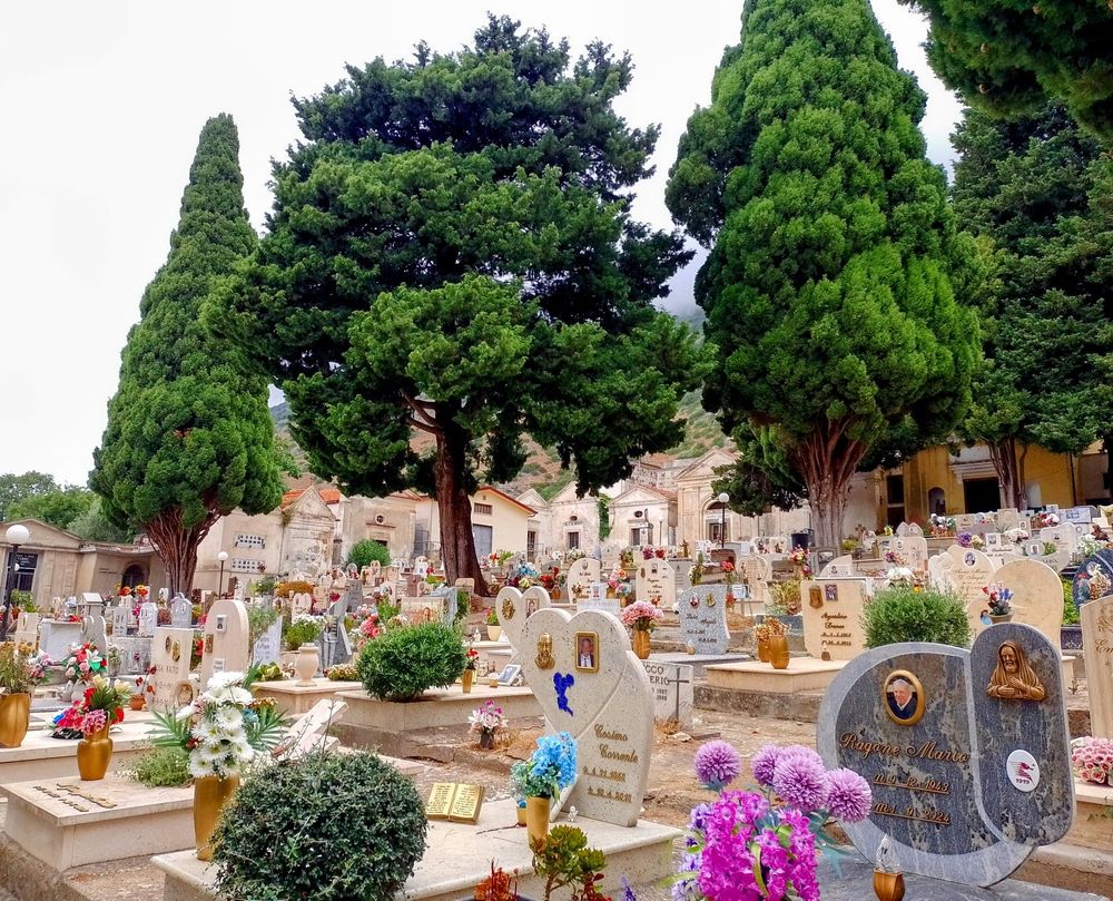 Photo of a small town cemetery. In the foreground there's a graveyard, with ground tombs, tombstones of various shapes and flowers. Amongst those there are some tall trees. In the background, two sides of the graveyard are lined with small chapels.