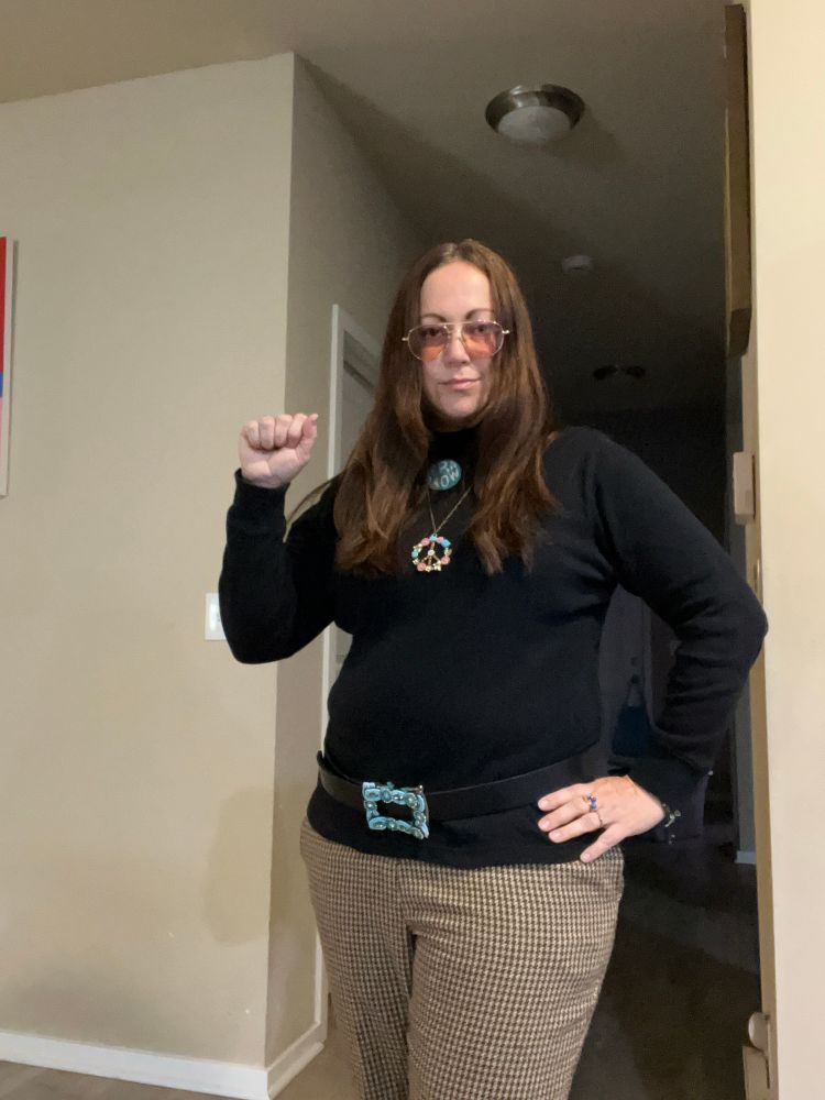 Woman in costume with checkered bell bottoms, hip hugger belt, turtleneck, peace pendant necklace, ERA now pin, and aviator glasses worn over center parted hair