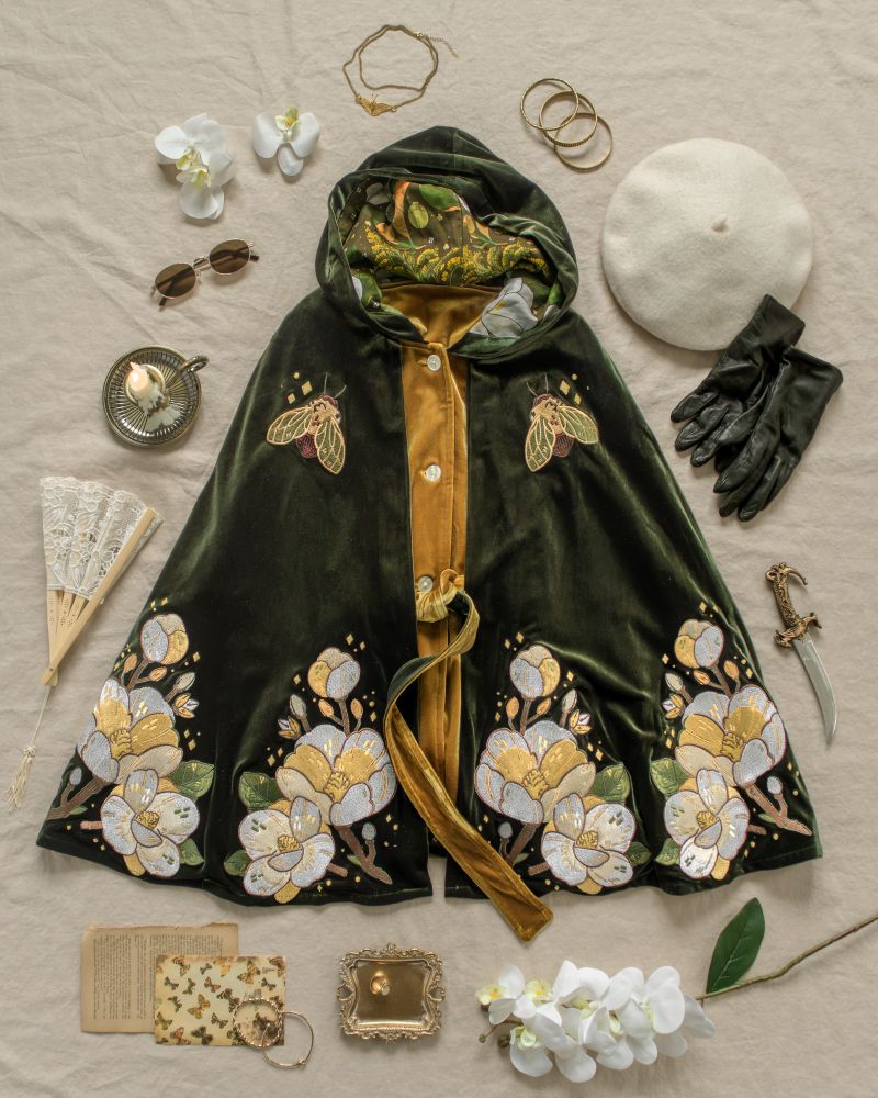 a flatlay against a cream background of a dark green velvet cloak with embroidery of magnolia flowers. 