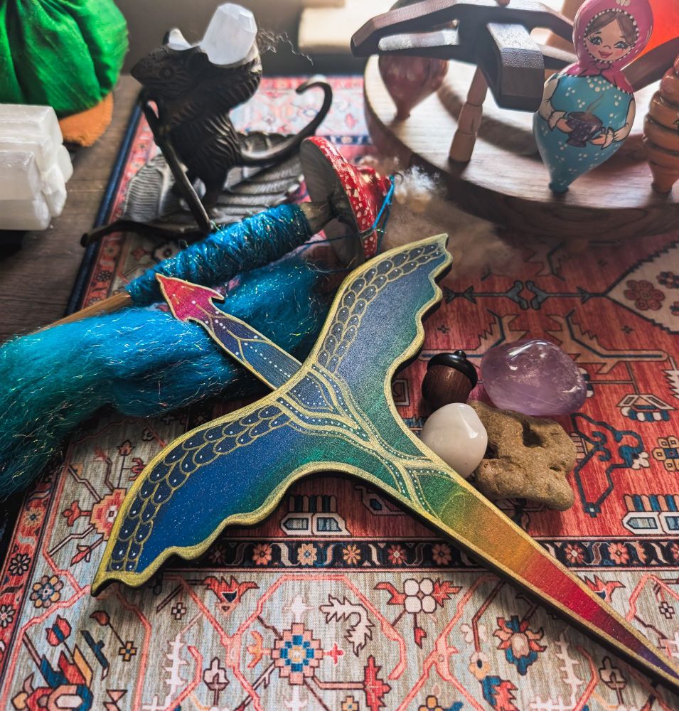 A rainbow and gold plying tool in the shape of a dragon sits on a kilim style desk mat. It rests on a tuft of sparkly blue fiber. Visible are some crystals, a hag stone, and acorn. Also visible are a number of spindles and Bernard the intrepid mouse statue that is actually a candleholder.
